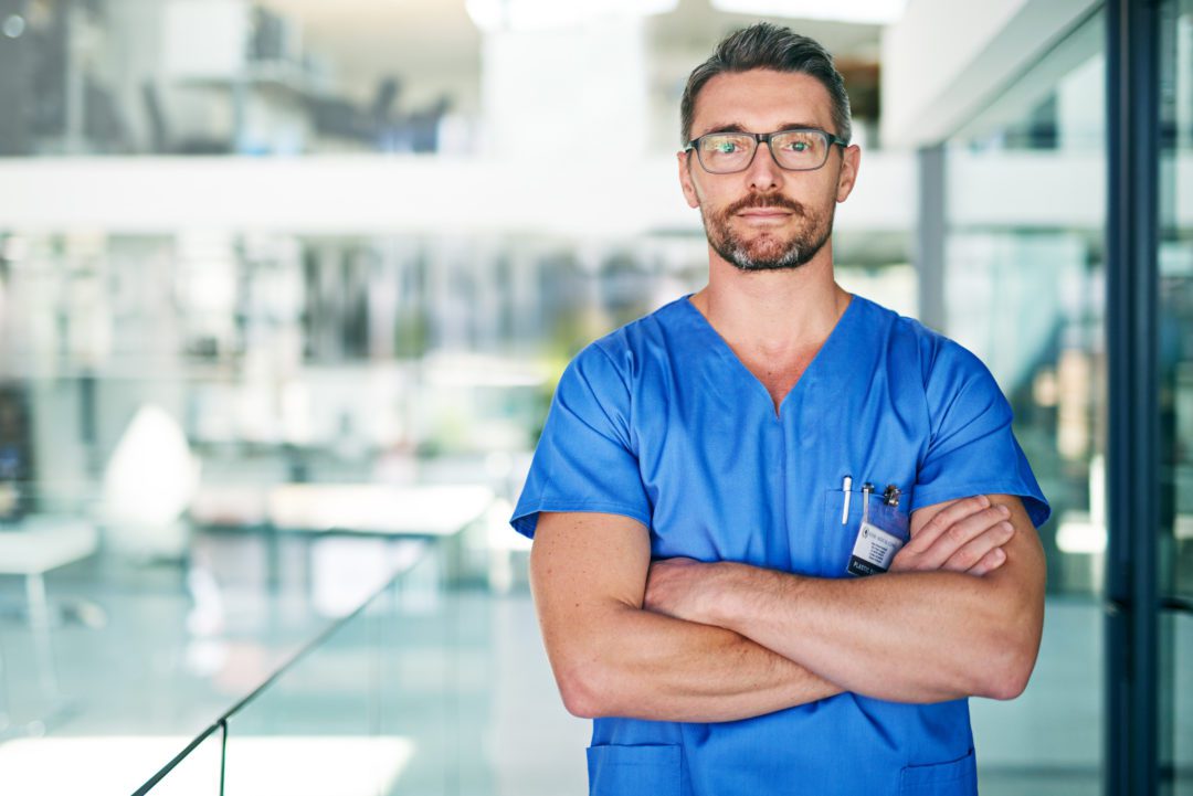 Male nurse with glasses and crossed arms looks directly at the camera