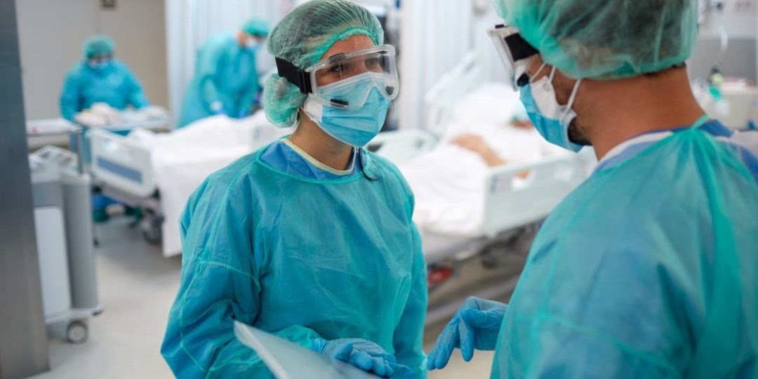 Two healthcare workers in PPE talking in a hospital room with patient and coworkers in the background