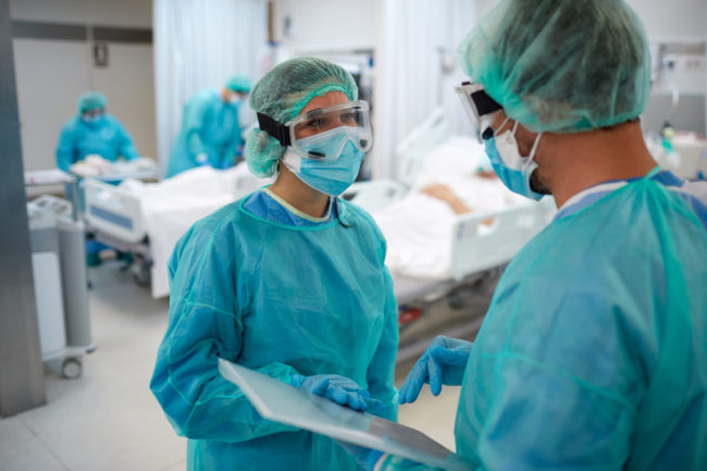 Two healthcare workers in PPE talking in a hospital room with patient and coworkers in the background