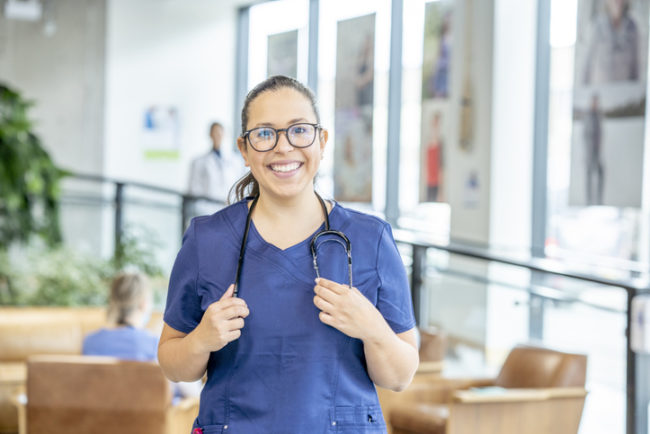 A nurse wearing blue scrubs smiles confidently at the camera. She is alone, but there are doctors and nurses walking around in the clinical space behind her.