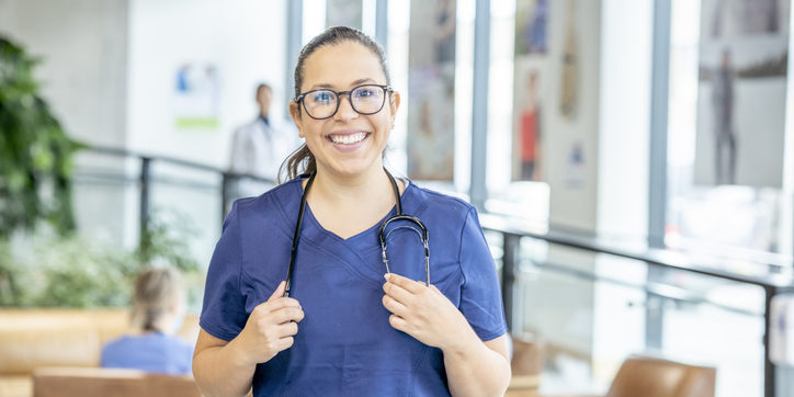 A nurse wearing blue scrubs smiles confidently at the camera. She is alone, but there are doctors and nurses walking around in the clinical space behind her.