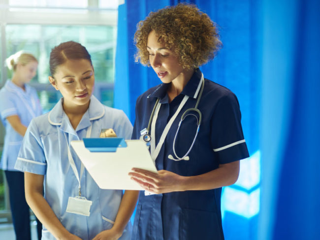 a nursing supervisor with a young ward nurse standing at the foot of a hospital bed chatting about the medical chart that she is holding . In the background a young nurse checks the dressing pack that she has got out of the trolley .