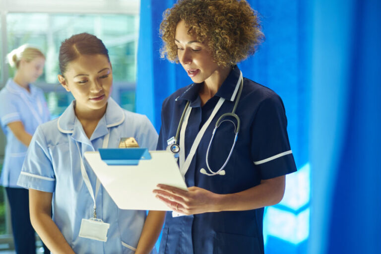 a nursing supervisor with a young ward nurse standing at the foot of a hospital bed chatting about the medical chart that she is holding . In the background a young nurse checks the dressing pack that she has got out of the trolley .