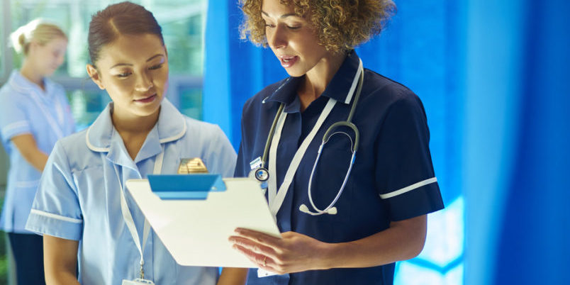 a nursing supervisor with a young ward nurse standing at the foot of a hospital bed chatting about the medical chart that she is holding . In the background a young nurse checks the dressing pack that she has got out of the trolley .